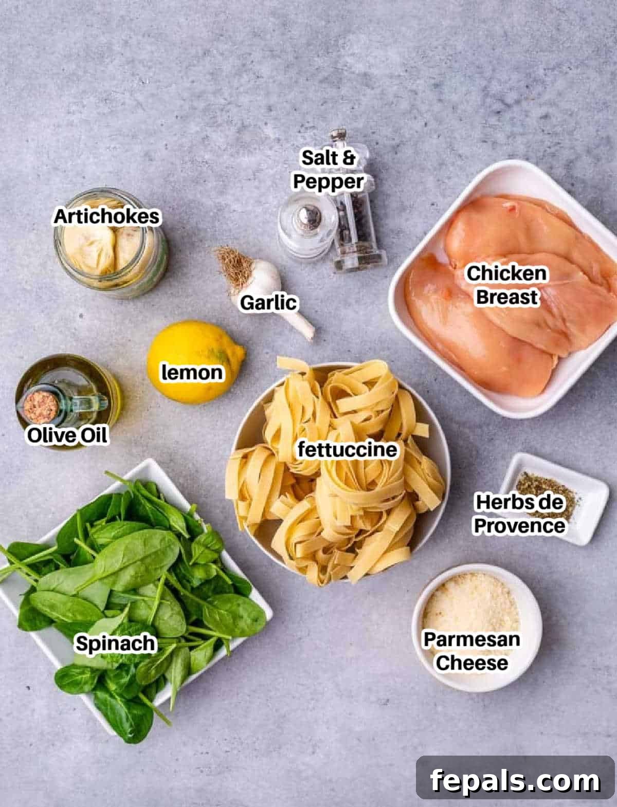 A flat lay of fresh ingredients including chicken breast, lemon, garlic, spinach, fettuccine pasta, artichoke hearts, olive oil, and parmesan cheese, ready to make lemon chicken pasta.