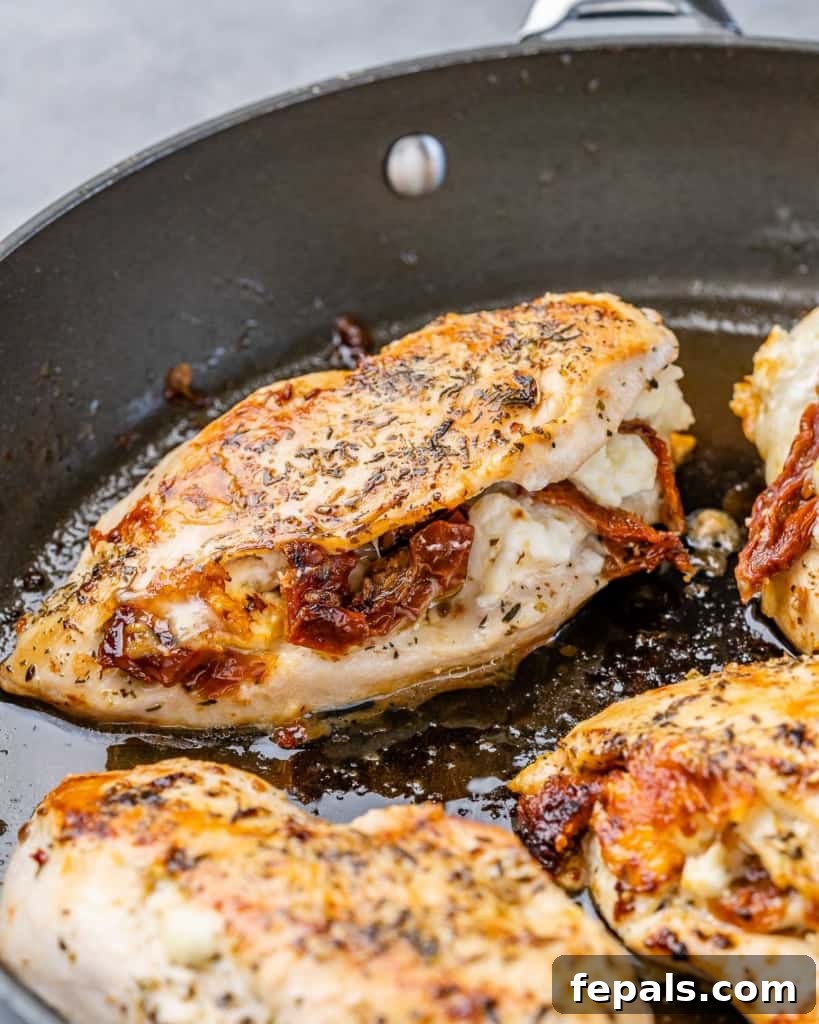 Savory Sun-dried Tomatoes and Cheese Stuffed Chicken 5 Close-up side shot of two golden brown stuffed chicken breasts, brimming with melted cheese and sun-dried tomatoes, resting in a rich sauce within a black skillet.