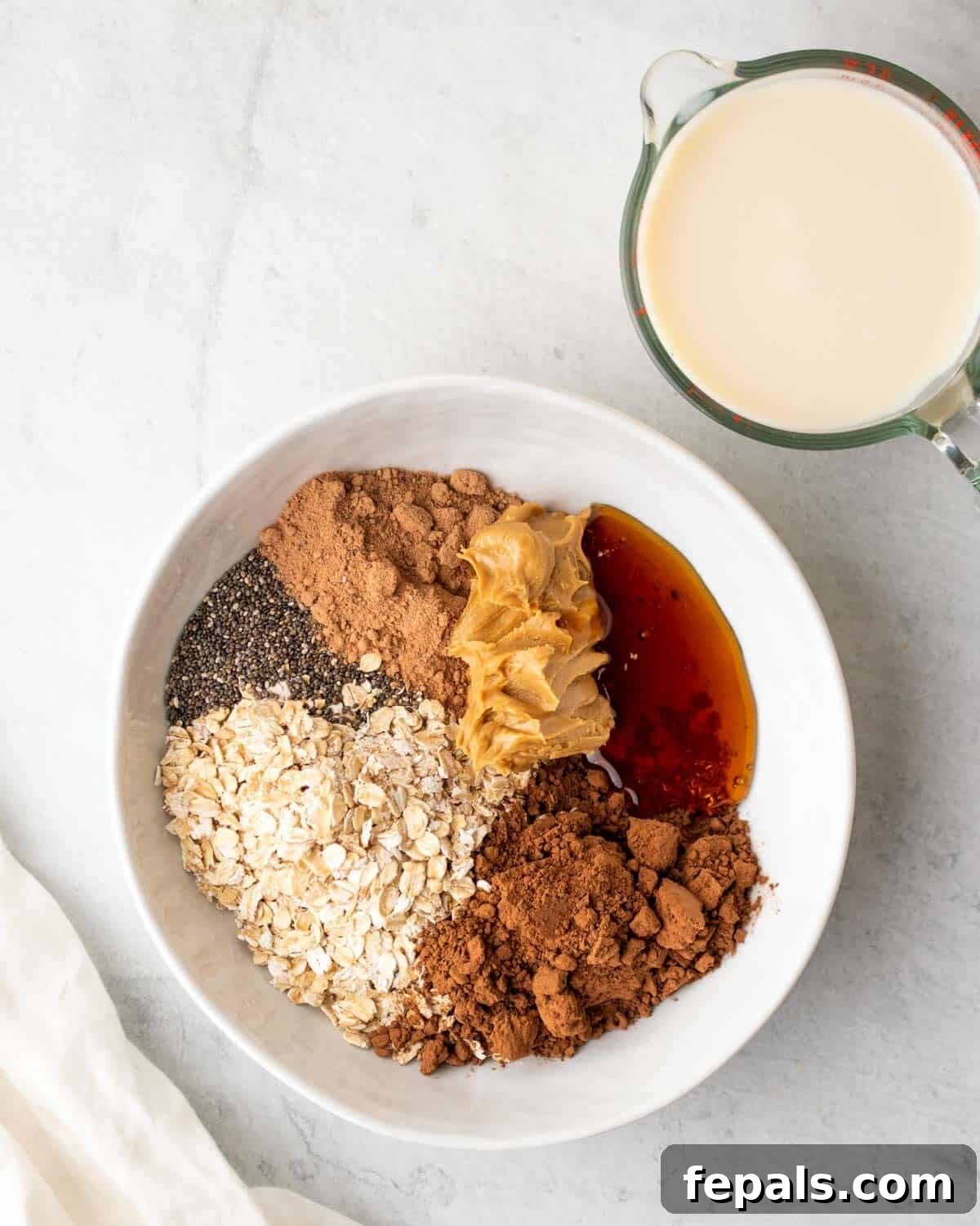 Dairy-free milk next to a bowl containing rolled oats, maple syrup, chia seeds, nut butter, cocoa powder, and protein powder