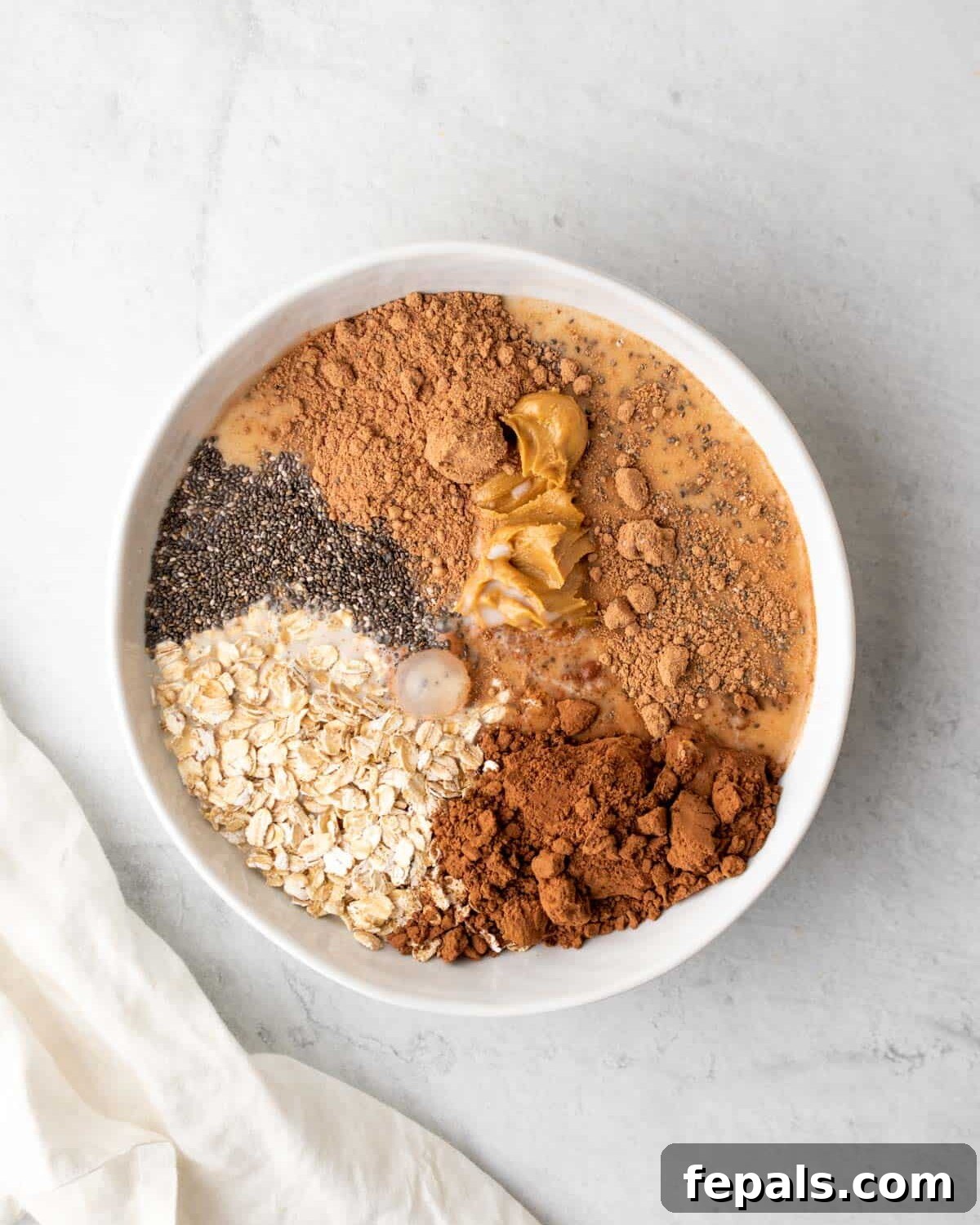 A bowl containing overnight oats mixture, maple syrup, chocolate powder, protein powder, and peanut butter, with milk being poured in
