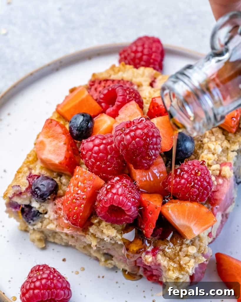 A golden-brown slice of baked oatmeal, artfully topped with a sprinkle of fresh berries, receiving a generous drizzle of rich, amber maple syrup, enhancing its delicious appeal.