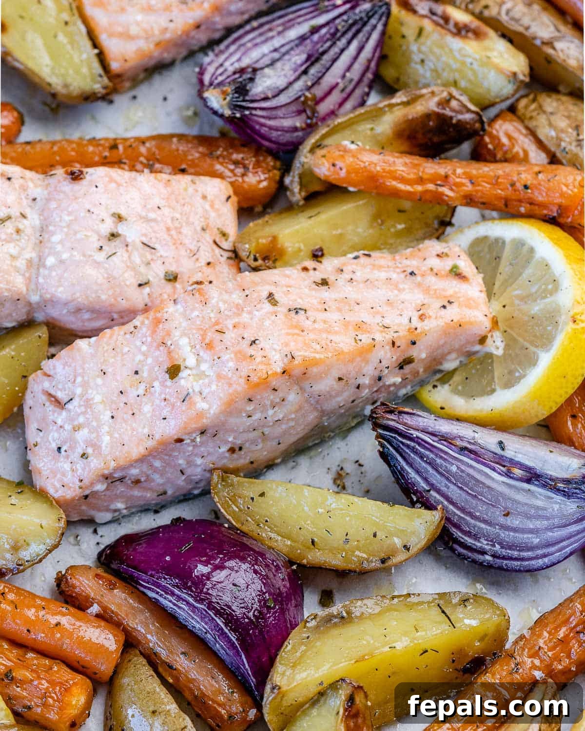 Close-up view of perfectly cooked salmon fillets with charred lemon slices, alongside golden roasted potatoes and tender root vegetables on a baking sheet.