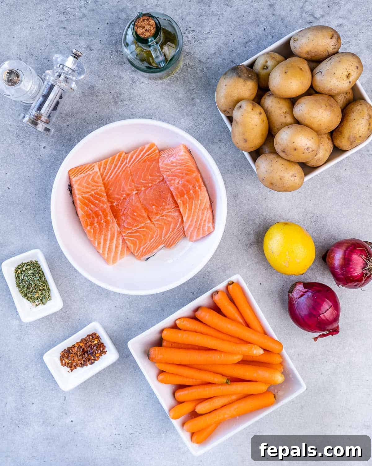 Selection of fresh ingredients laid out: baby potatoes, olive oil, salt, pepper, salmon fillets, Italian seasoning, chili flakes, carrots, two red onions, and a lemon.