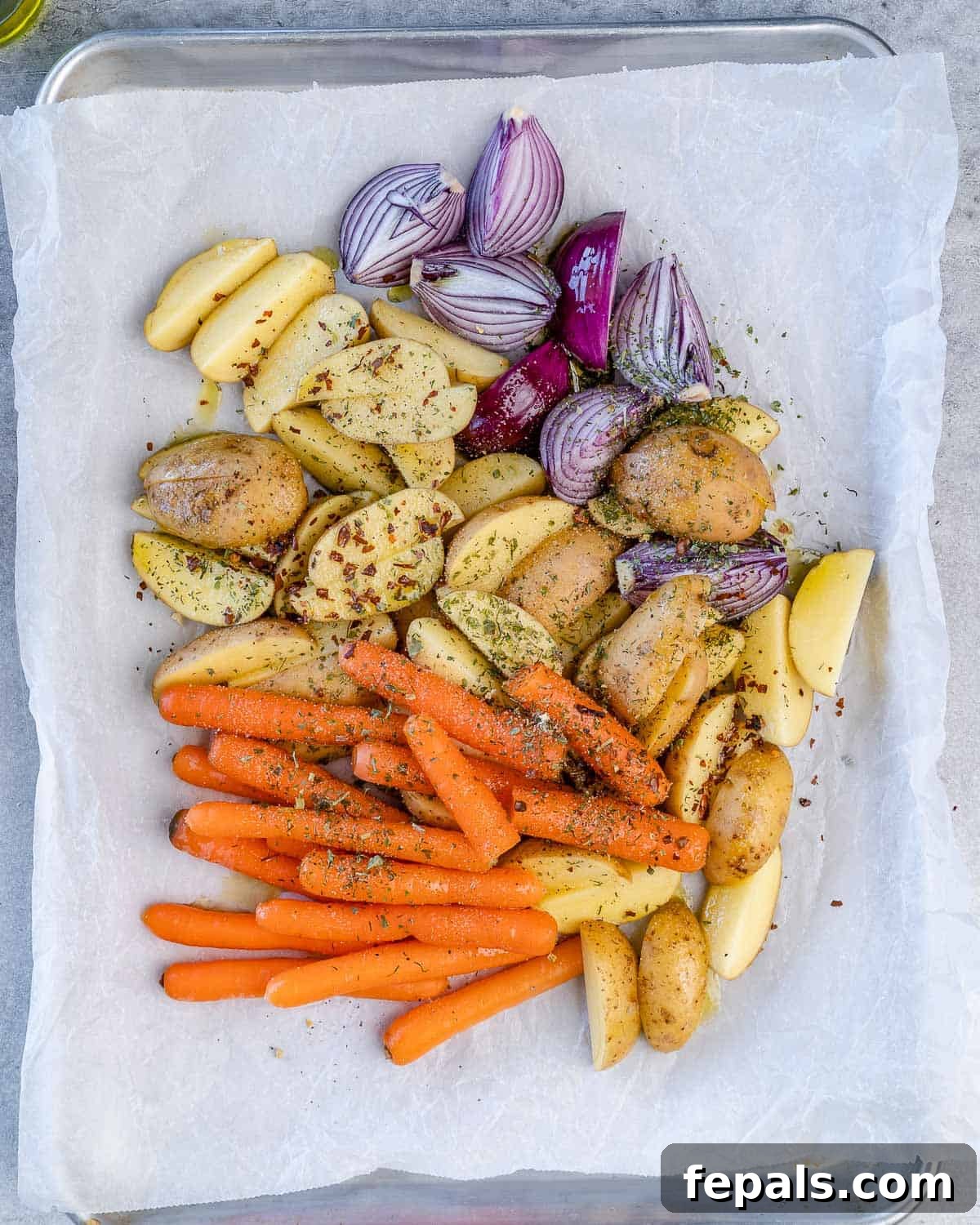 Assortment of red onions, carrots, and potatoes arranged on a baking sheet with parchment paper, generously topped with a blend of seasonings.
