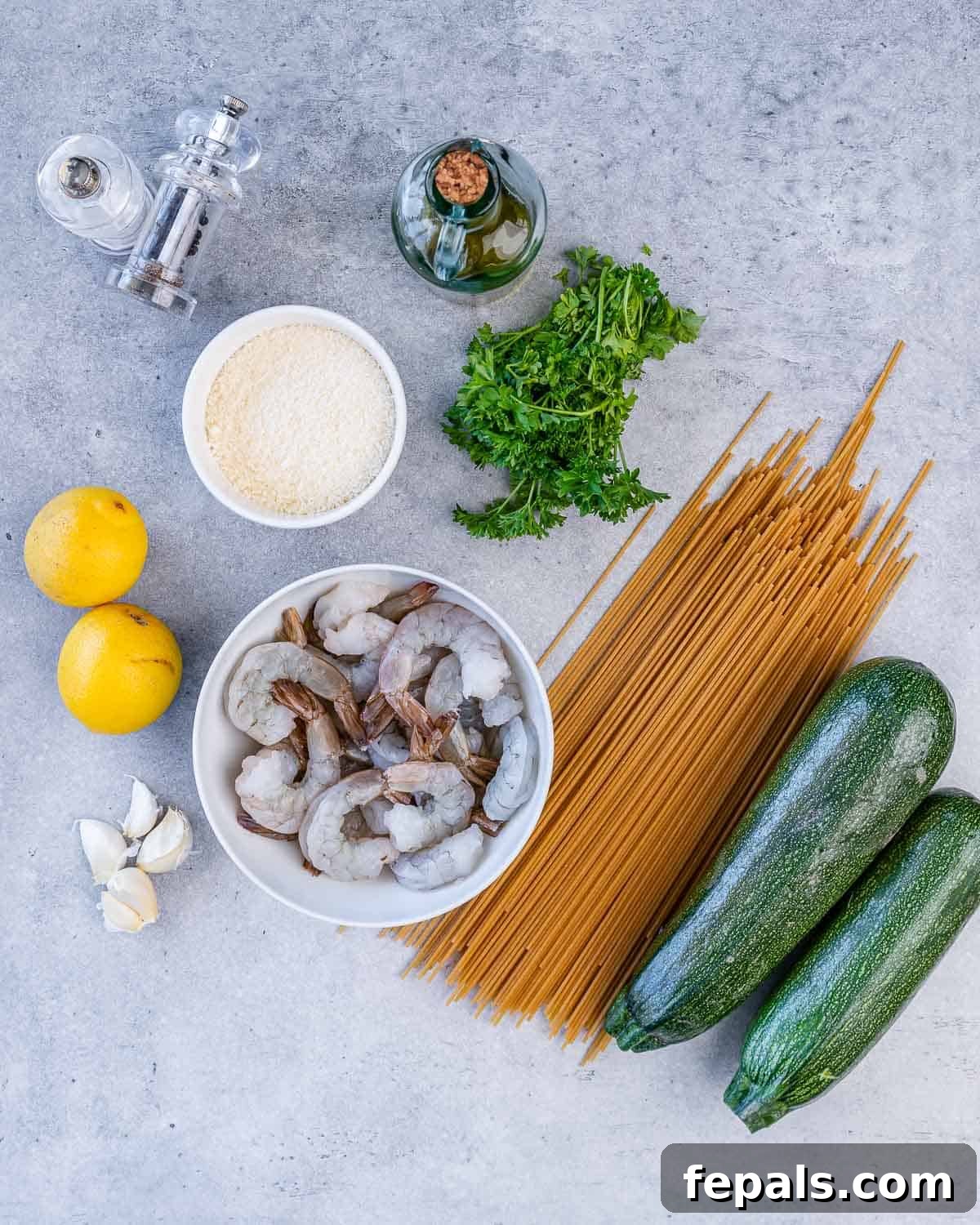 Zesty Lemon Shrimp Pasta with Zucchini Noodles 4 A flat lay arrangement of fresh ingredients for lemon shrimp pasta: uncooked spaghetti, two zucchini, raw shrimp, parsley, olive oil, parmesan cheese, salt, pepper, two lemons, and garlic cloves.