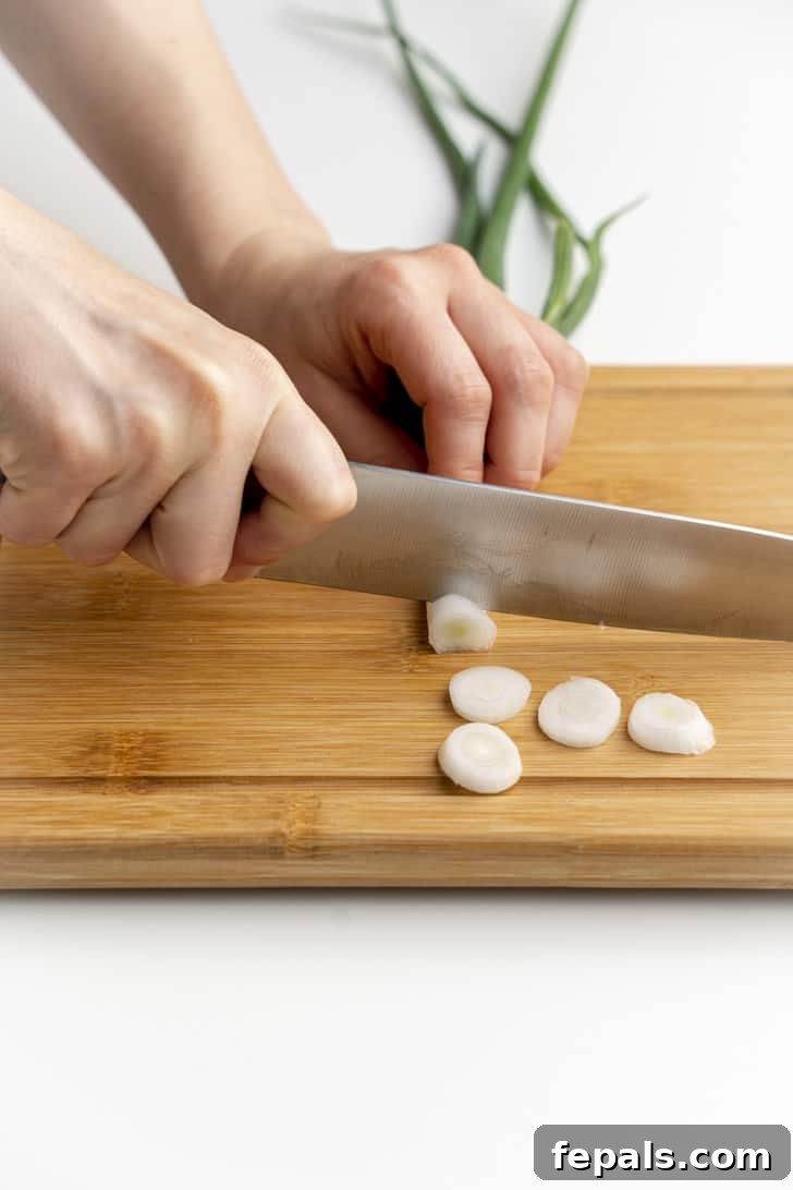 A sharp knife slicing through a single green onion on a wooden cutting board, creating even rounds