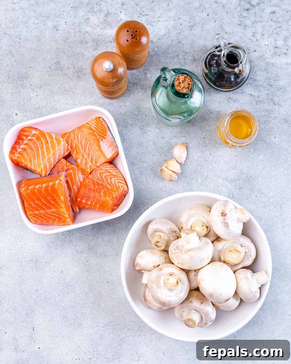 Arrangement of fresh ingredients: salmon fillets, whole button mushrooms, garlic cloves, honey, balsamic vinegar, salt, and pepper on a gray kitchen surface, ready for cooking.