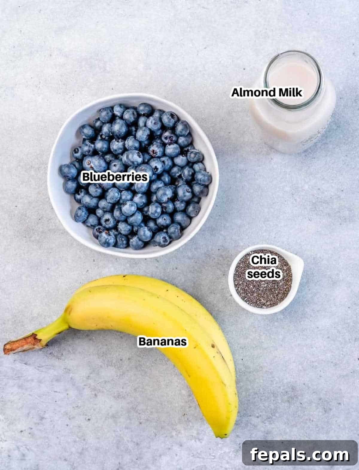 A flat lay image displaying the key ingredients needed to make a blueberry banana smoothie: fresh blueberries, ripe bananas, a bottle of milk, and a small bowl of chia seeds.