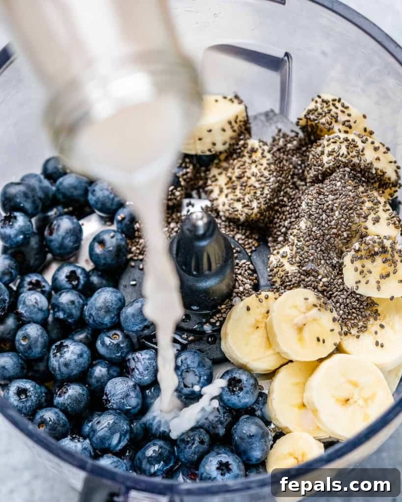 Pouring milk into a high-speed blender already filled with fresh blueberries, sliced bananas, and a spoonful of chia seeds, illustrating the first step of making the smoothie.