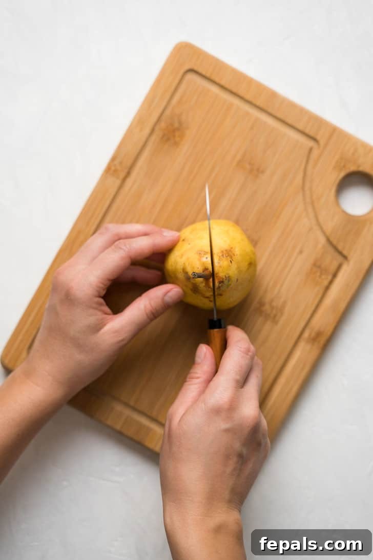 How to Cut a Pear Into Cubes A hand holds a pear firmly on a wooden cutting board as a sharp knife begins to slice through its center.