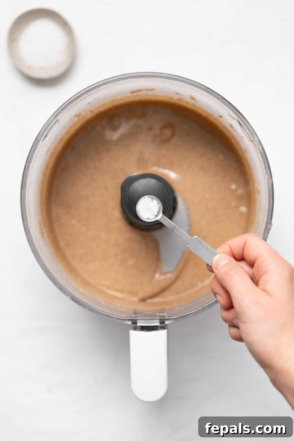 A hand adding salt to the creamy almond butter swirling in the food processor.