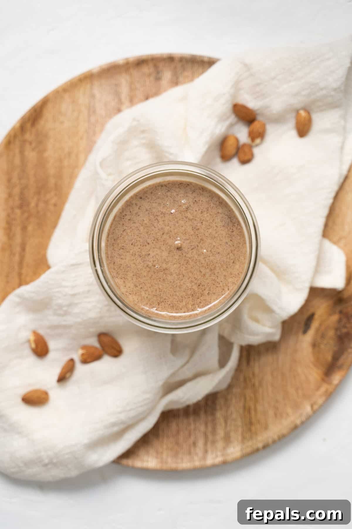 Freshly made almond butter poured into a clear glass jar, set against a white linen backdrop.