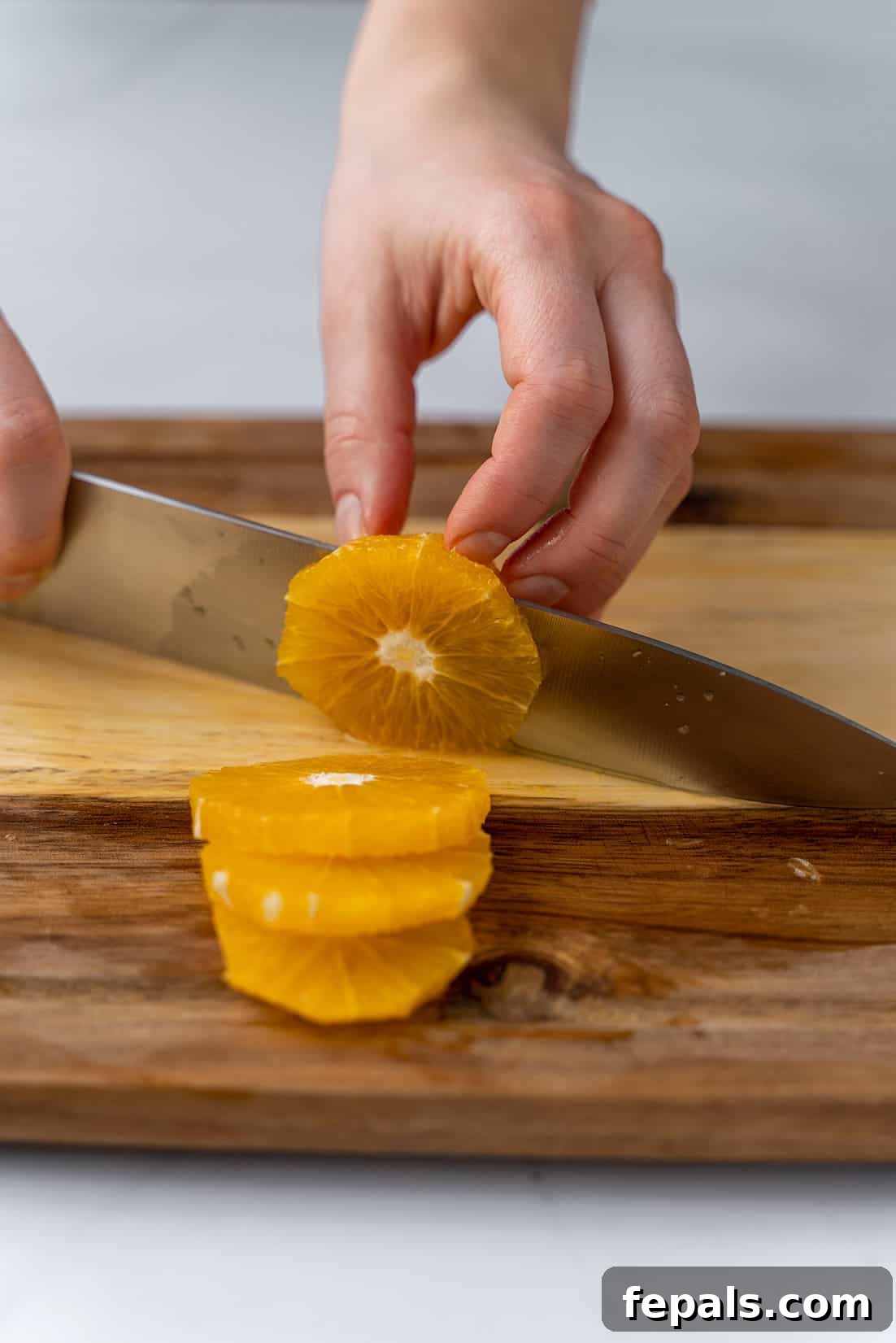 A hand gently slicing an orange into perfect rings with a knife on a wooden cutting board.