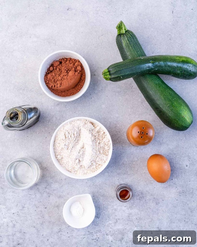 Two fresh zucchini next to bowls containing ingredients like cocoa powder, flour, and a measuring cup of maple syrup, all arranged on a wooden surface.