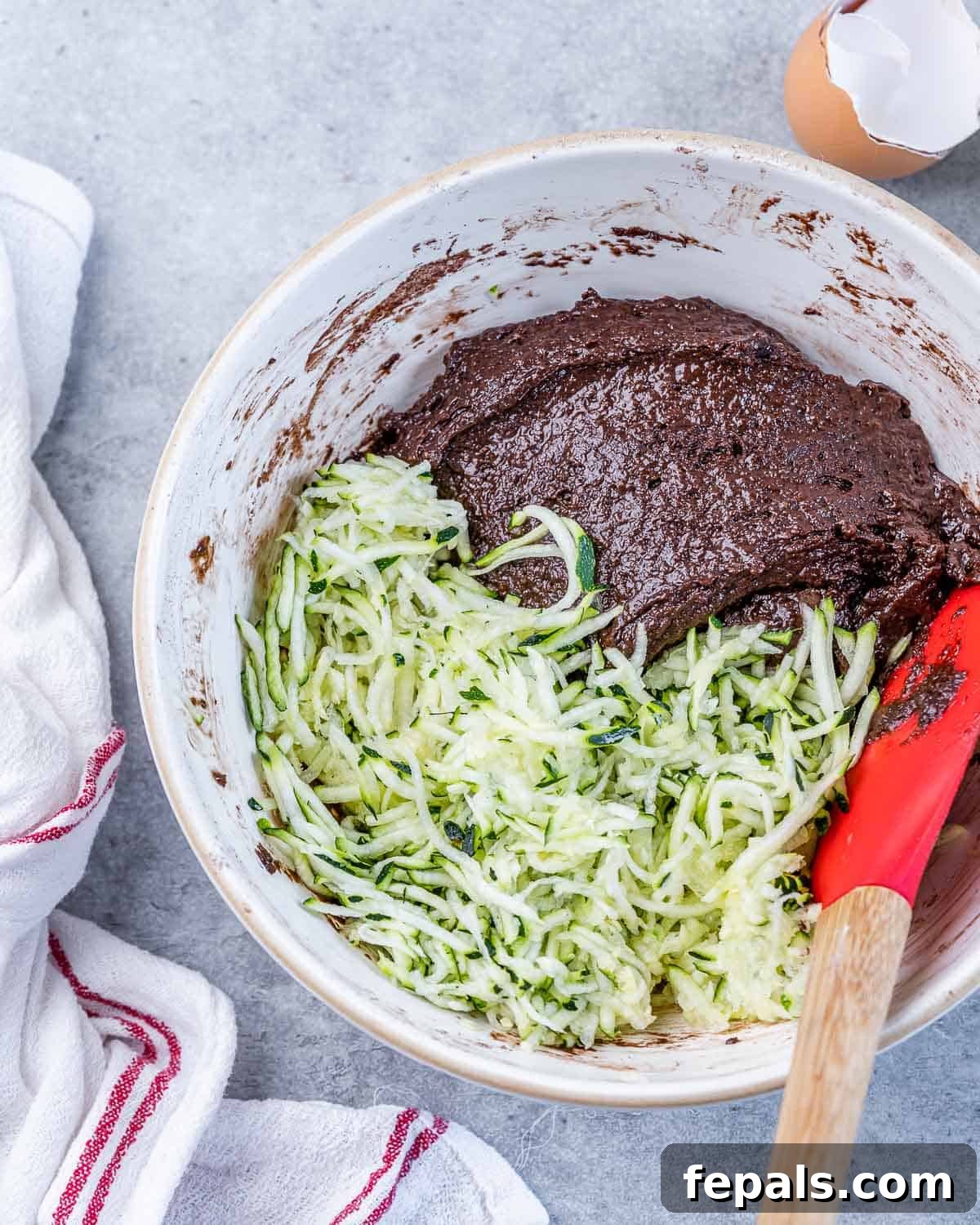 A close-up shot of shredded zucchini being added to a bowl of rich chocolate brownie batter.