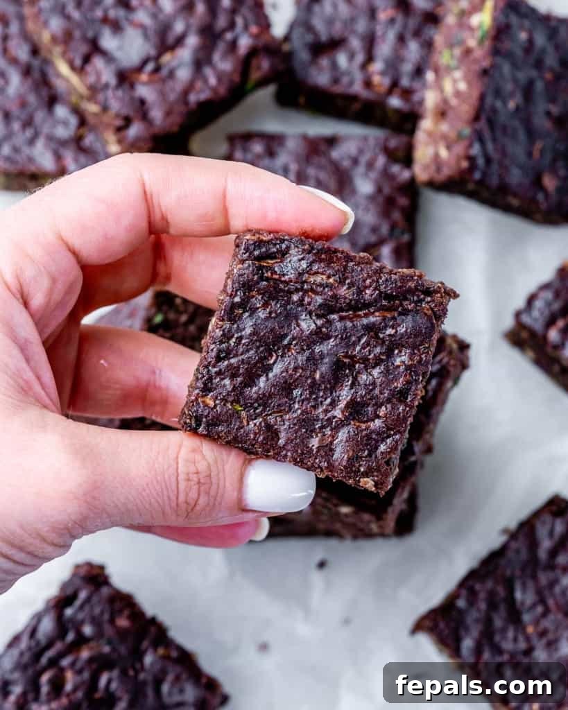A woman's hand holding a single square of a healthy zucchini brownie, showcasing its rich chocolate color and dense texture.