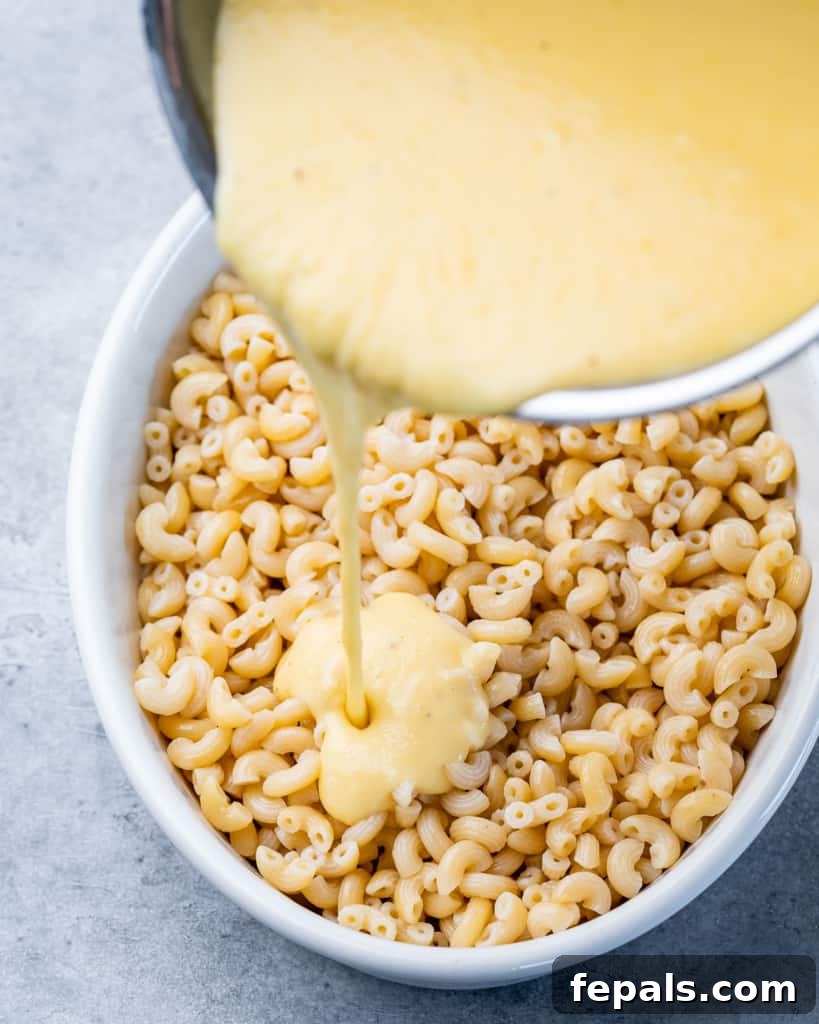 Cheese sauce being poured over cooked elbow noodles in a white baking dish.
