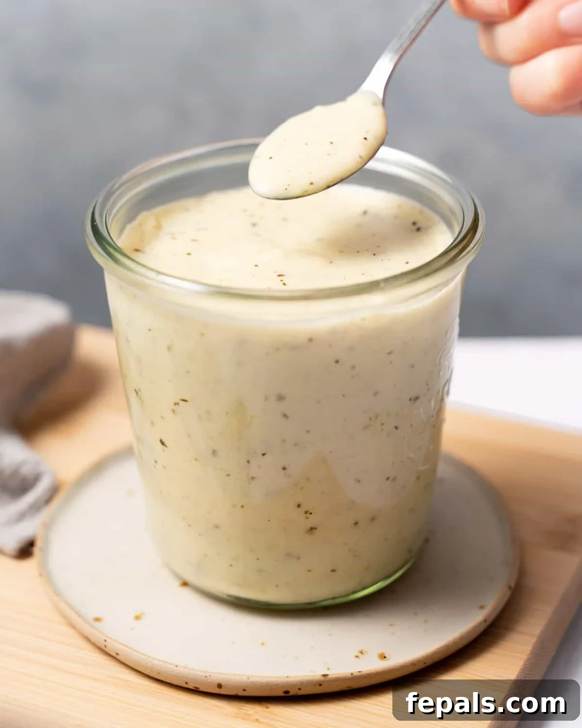 Woman's hand spooning Healthy Alfredo Sauce from a glass jar, highlighting its creamy texture.