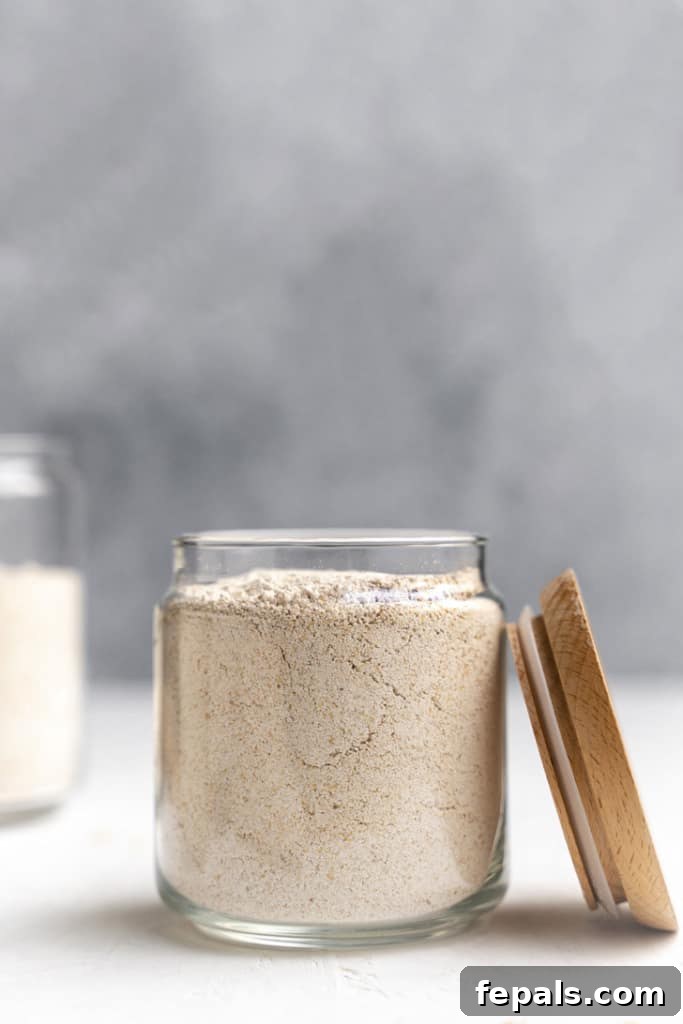 A glass jar filled with fine oat flour, positioned beside its lid, showcasing the smooth texture of the homemade flour.
