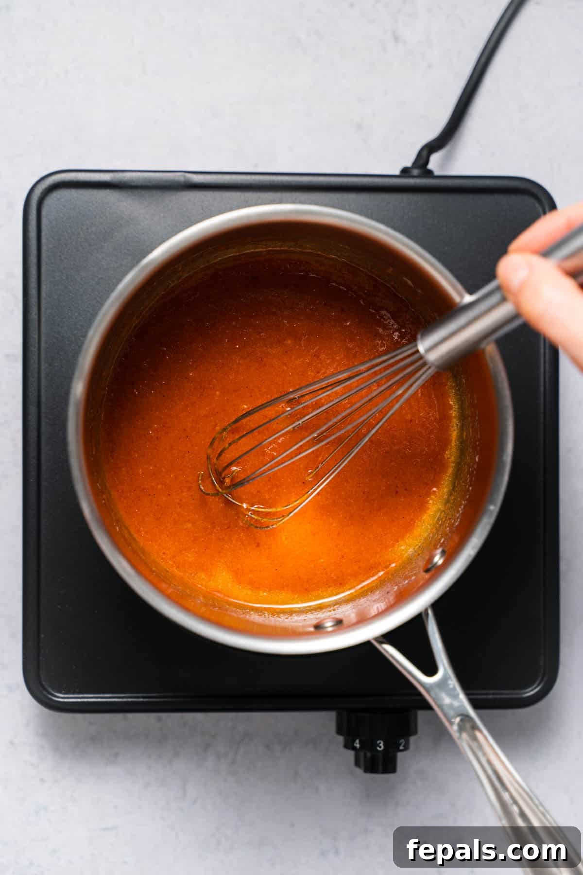 A whisk vigorously mixing the buffalo sauce ingredients in a saucepan, creating a smooth, emulsified, orange mixture.