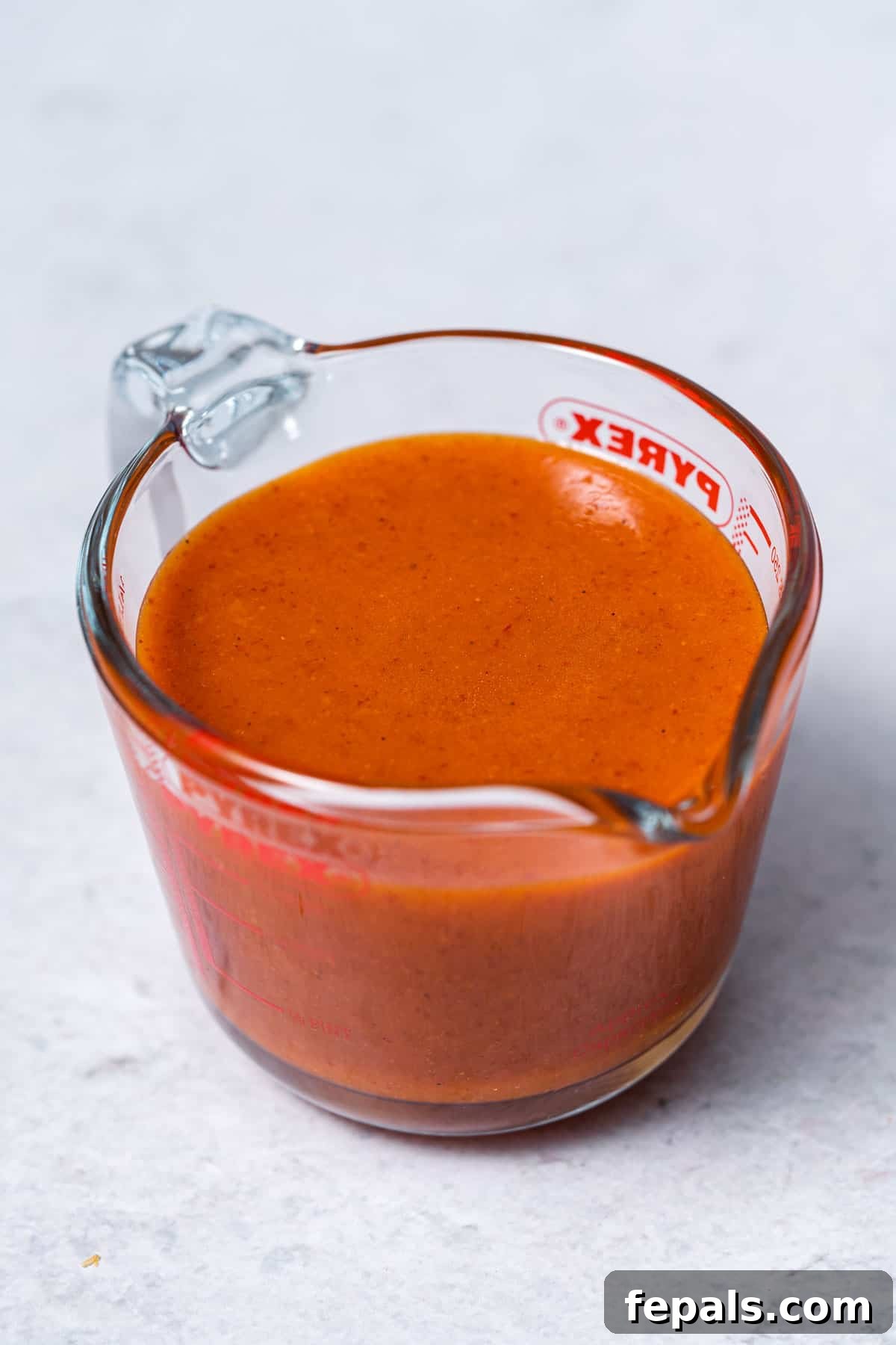 A clear glass measuring cup filled with vibrant, smooth homemade buffalo sauce, ready to be poured or served.