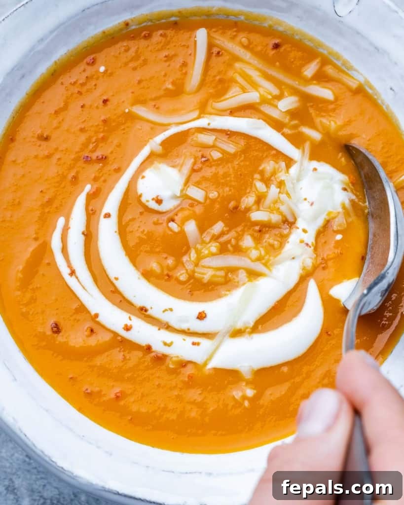 Top view close up of butternut squash soup in a white bowl with a spoon, topped with a sour cream swirl and fresh herbs.