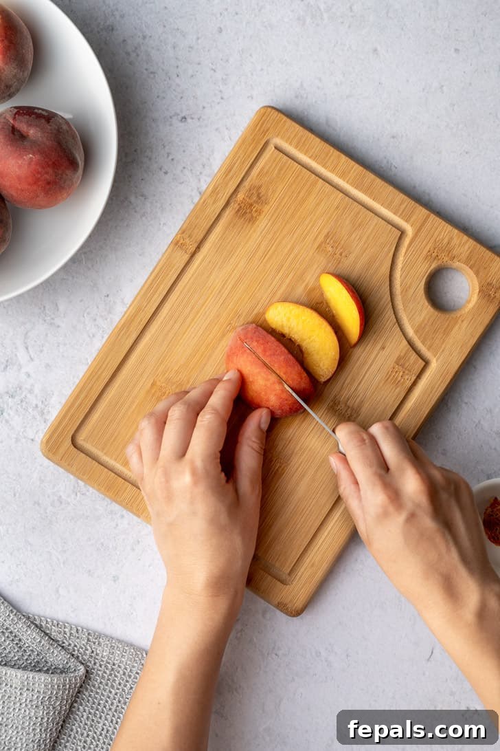A hand holds a knife, carefully continuing to slice through a peach half, emphasizing the method for consistent peach slices.