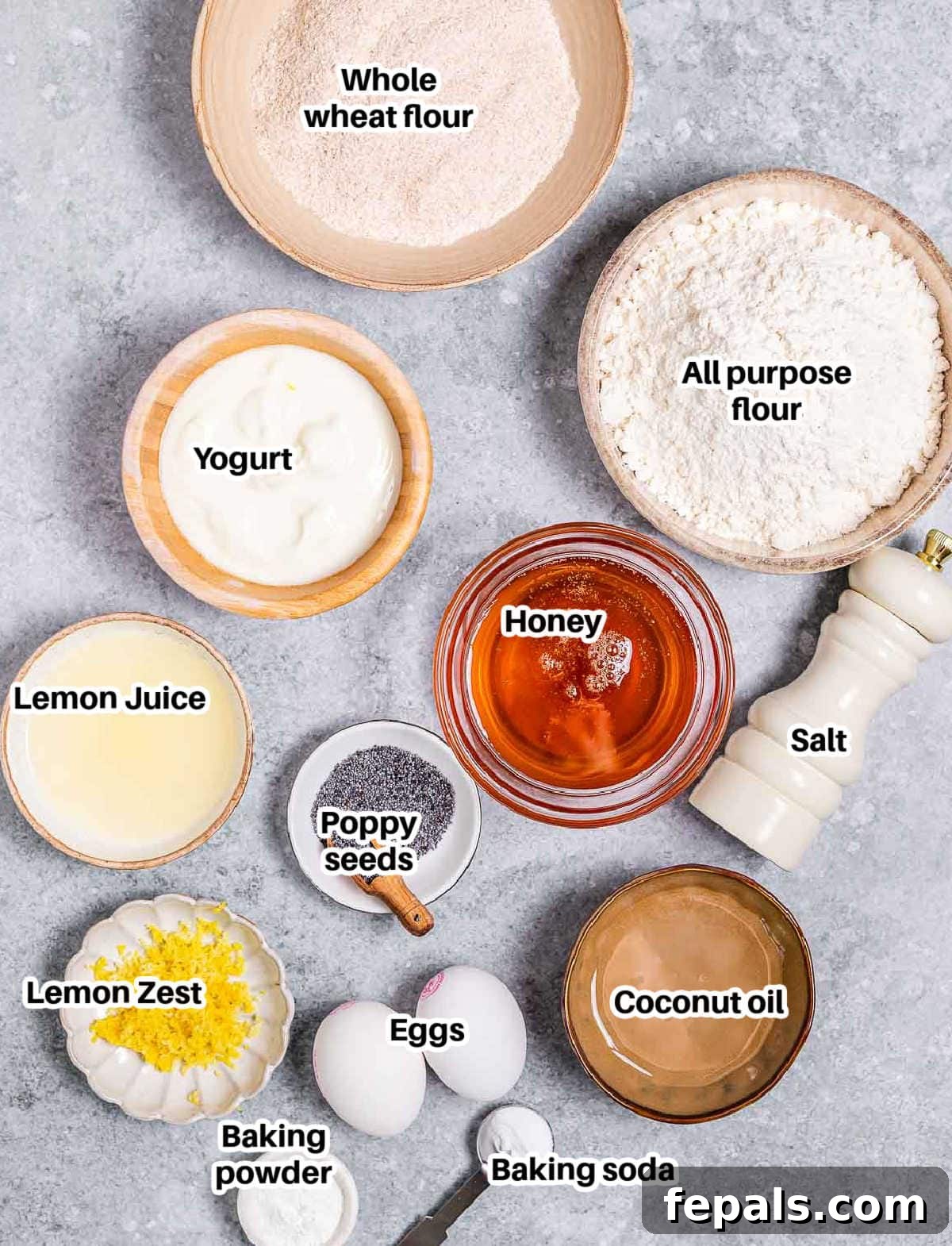 A curated display of fresh ingredients for lemon poppy seed muffins, including whole wheat flour, all-purpose flour, lemons, poppy seeds, eggs, yogurt, honey, and baking essentials, laid out on a rustic surface.