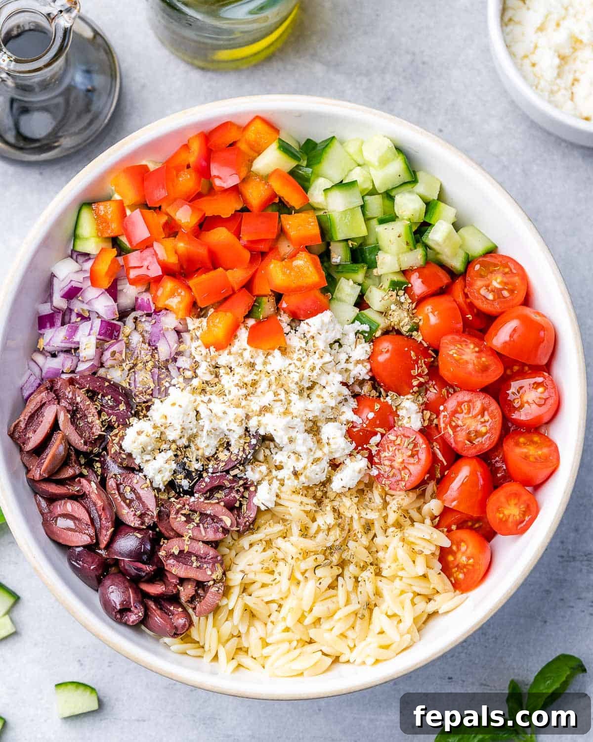 Zesty Greek Orzo Salad 7 Cooked orzo pasta being added to a large serving bowl with chopped vegetables.