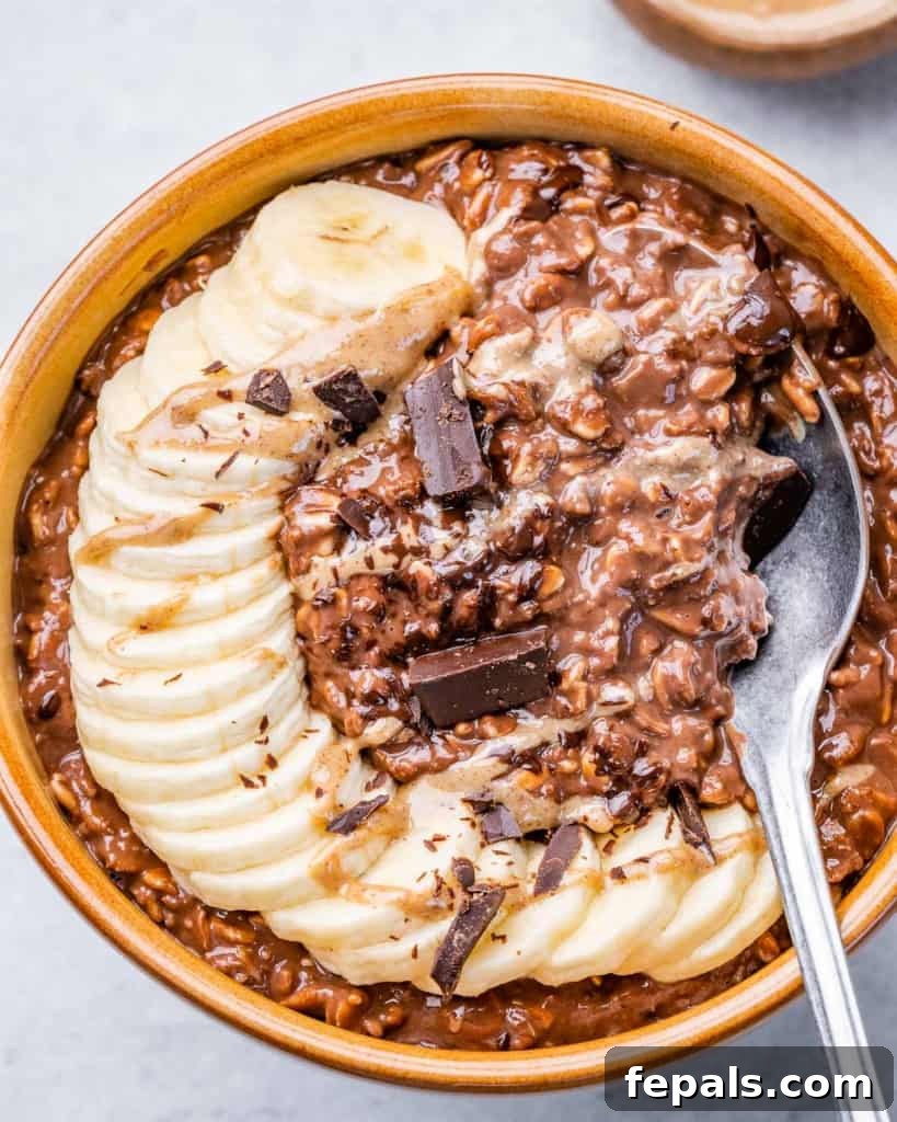 top view of chocolate oatmeal in a brown bowl with sliced bananas and shaved chocolate, spoon in the bowl of oatmeal