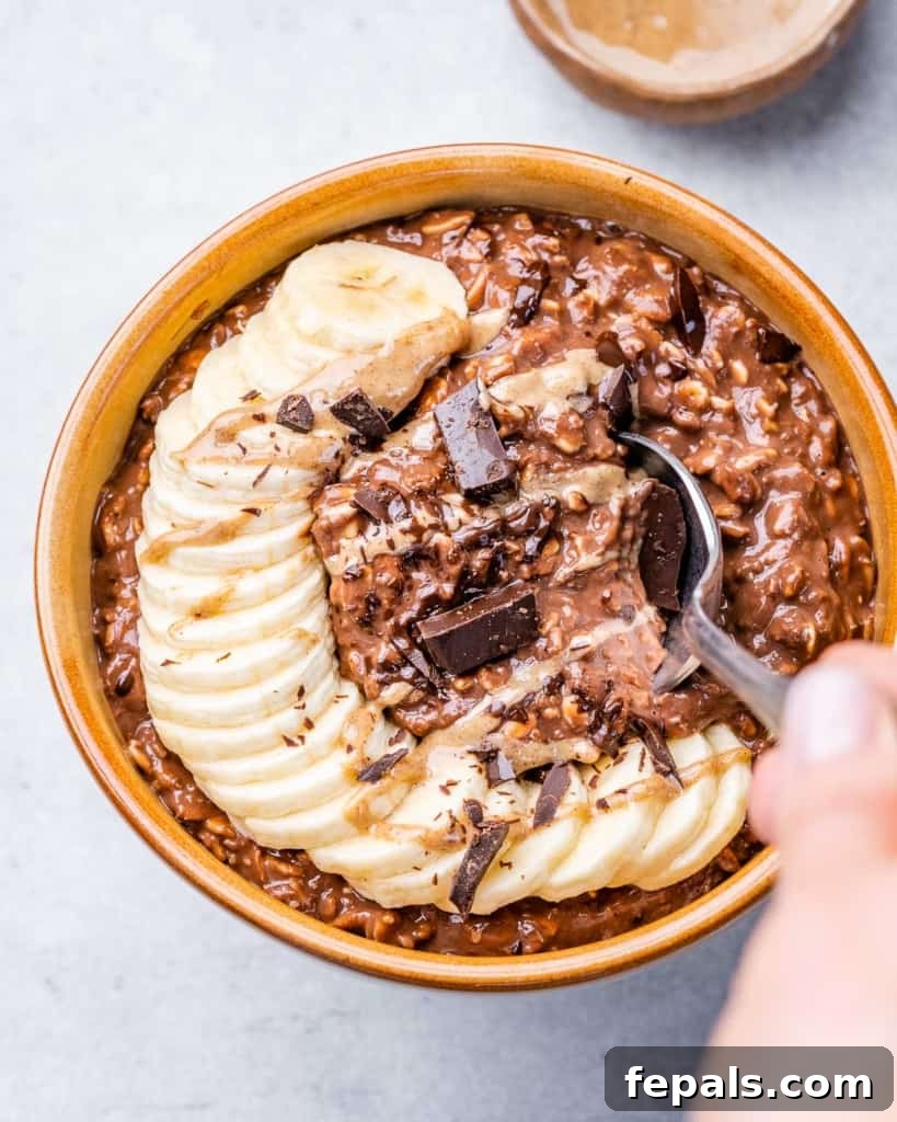 hand holding a spoon over chocolate oatmeal bowl topped with sliced bananas and shaved chocolate