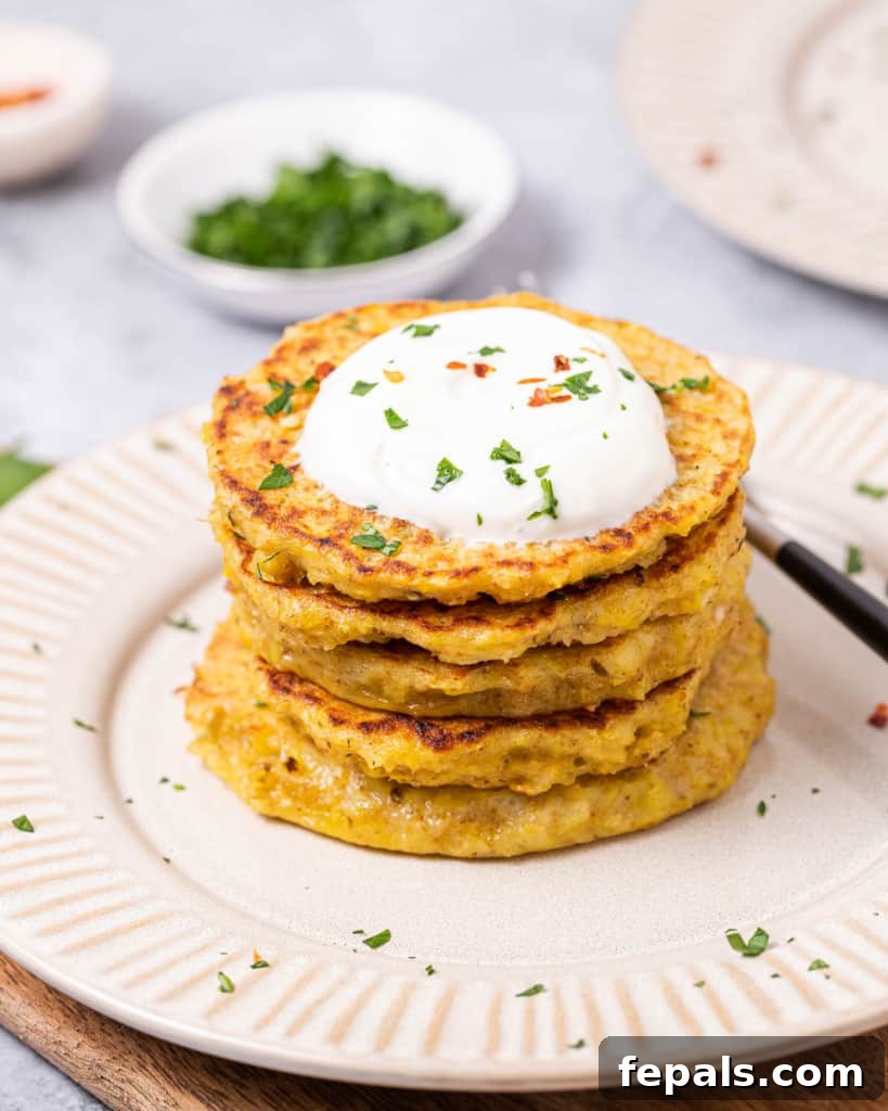 Timeless Golden Potato Fritters 10 Side shot of potato pancakes stacked on each other on a white plate, topped with yogurt and a sprig of fresh herb.