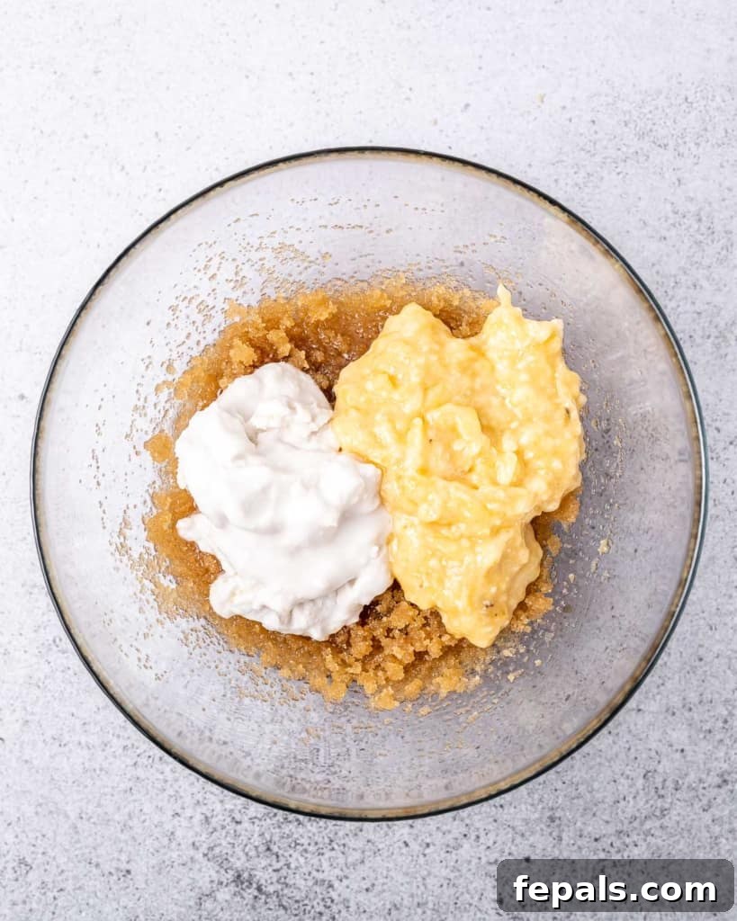 A clear bowl containing a mixture of mashed banana, coconut cream, and sugar, blended smoothly and ready for the next wet ingredients.