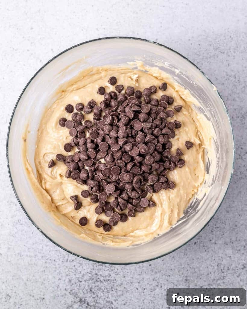 Vegan chocolate chips generously sprinkled over the thick cookie dough batter in a mixing bowl, ready to be gently folded in.