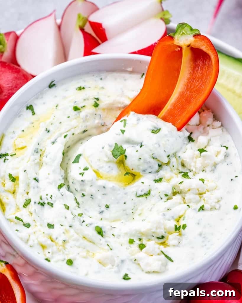 Red bell pepper slice being dipped into a white, creamy herbed whipped feta dip in a small bowl.
