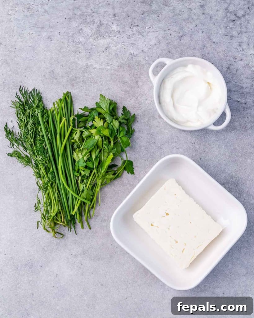 Ingredients for herbed whipped feta dip: a block of feta cheese, a bowl of Greek yogurt, and three small piles of fresh herbs (dill, parsley, chives) on a cutting board.