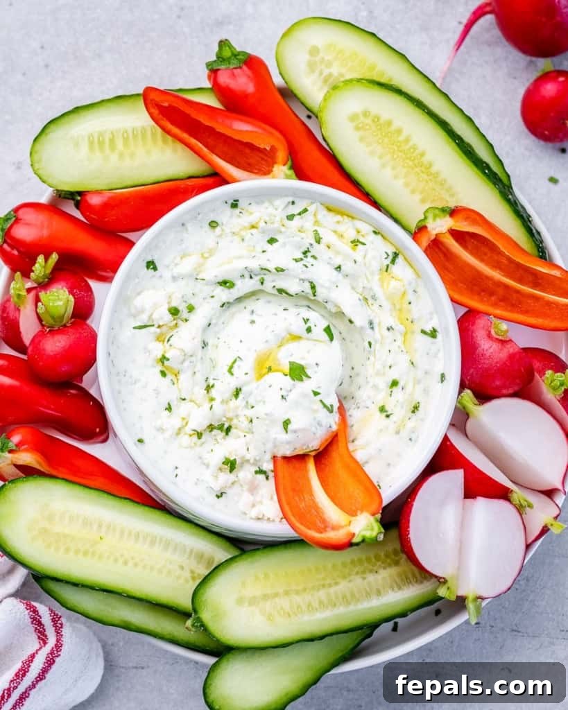 Top view of a beautifully presented herbed whipped feta dip in a white bowl, garnished with herbs and a swirl of olive oil, with an assortment of sliced fresh vegetables arranged elegantly on the side.