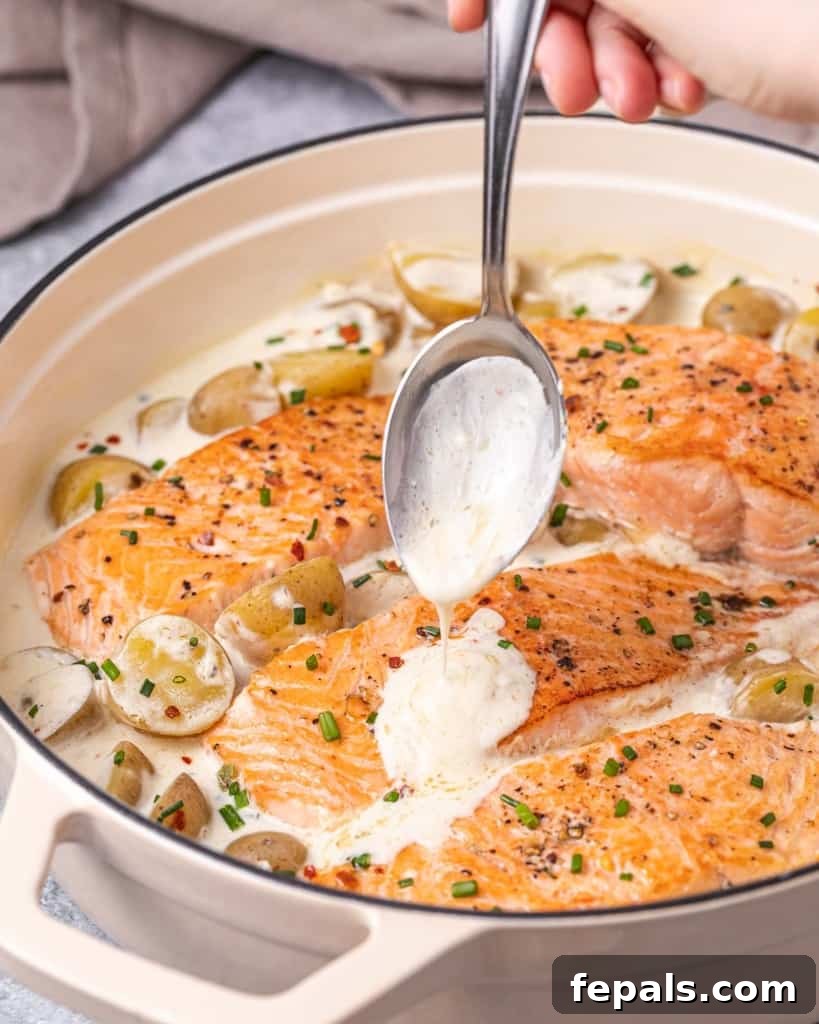 Hand holding a spoon, drizzling creamy white sauce over a perfectly cooked salmon fillet in a skillet.