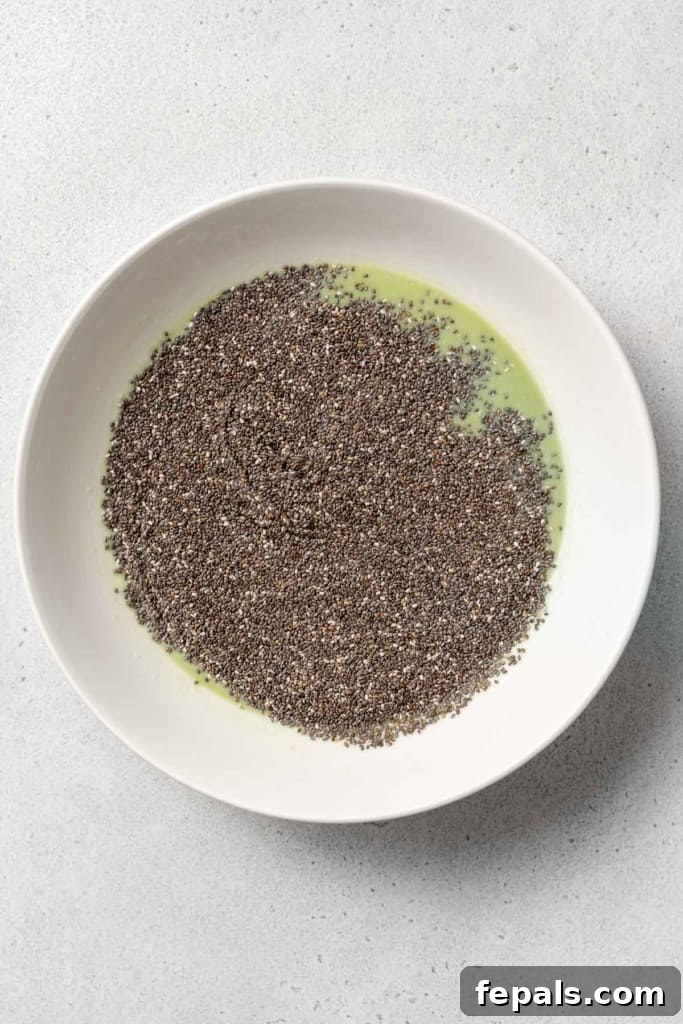 Small chia seeds are added to the vibrant green matcha milk mixture in a bowl, preparing for the gelling process.
