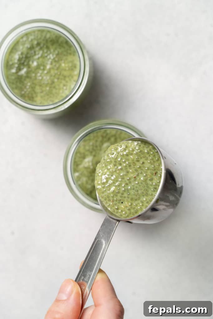A hand uses a measuring scoop to transfer the matcha chia pudding mixture into a glass jar, preparing it for chilling.