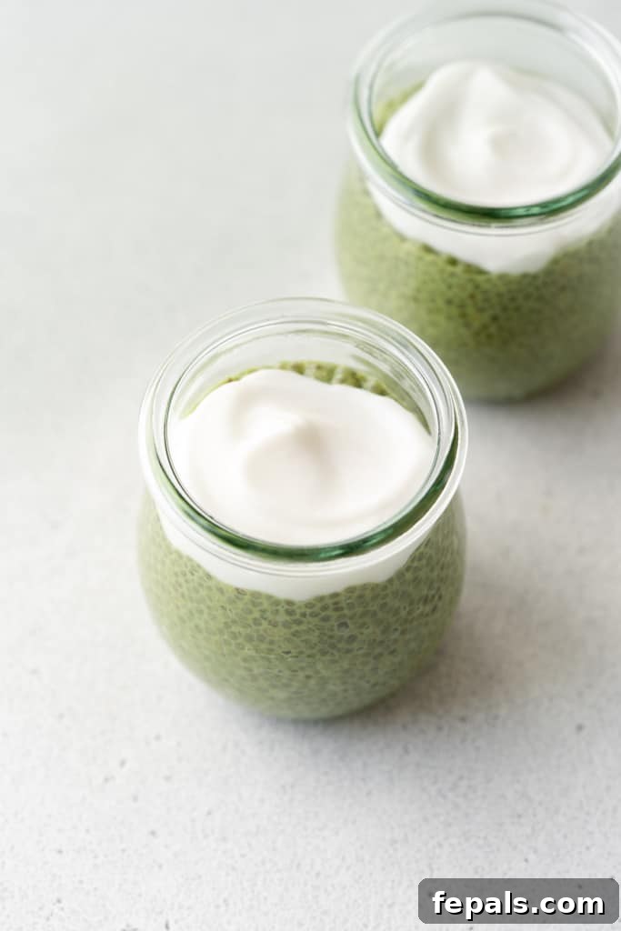 A spoonful of creamy coconut whip is added on top of the vibrant matcha chia pudding in a jar, preparing it for serving.