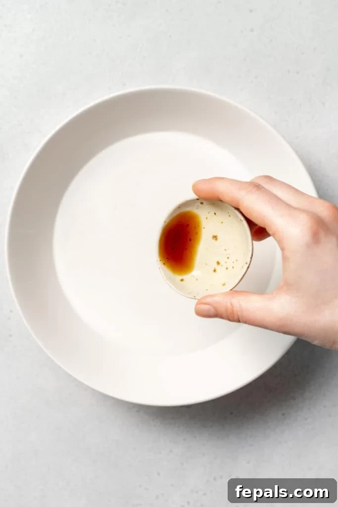 A small measuring spoon adds a splash of vanilla extract into the bowl containing milk, enhancing the pudding's flavor.