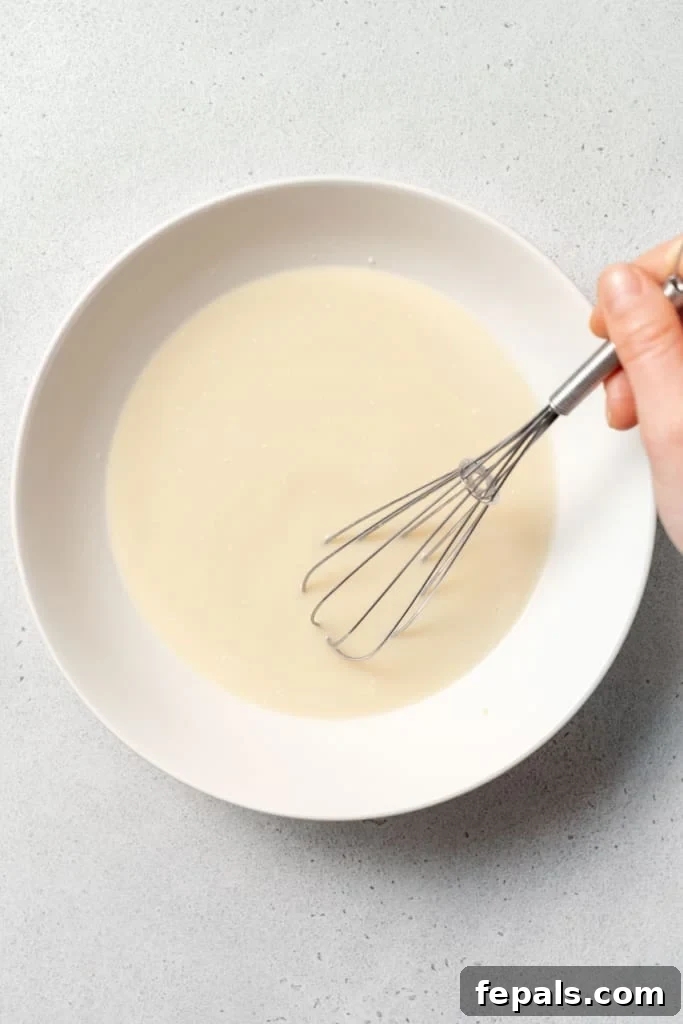 A whisk vigorously mixes the milk, vanilla, and sweetener in a bowl, ensuring a smooth and homogenous liquid base for the pudding.