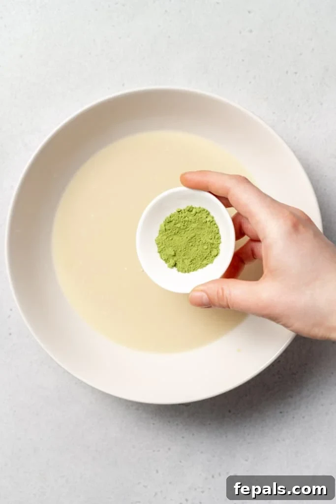 A hand sifts or sprinkles vibrant green matcha powder over the liquid mixture in the bowl, adding color and flavor.