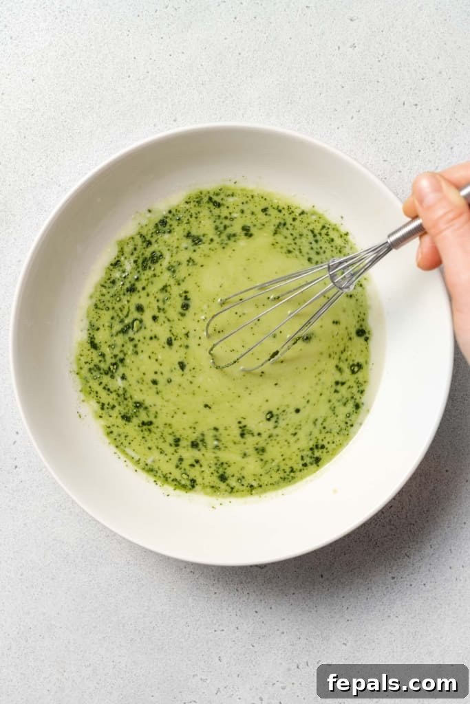A hand, holding a whisk, actively mixes the matcha powder into the milk, ensuring a smooth, lump-free consistency.