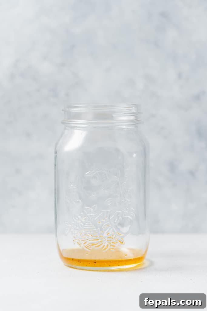 Refreshing Layered Chickpea Cucumber Jar 6 Pouring a golden salad dressing into the bottom of a clear mason jar, preparing it for layered salad ingredients.