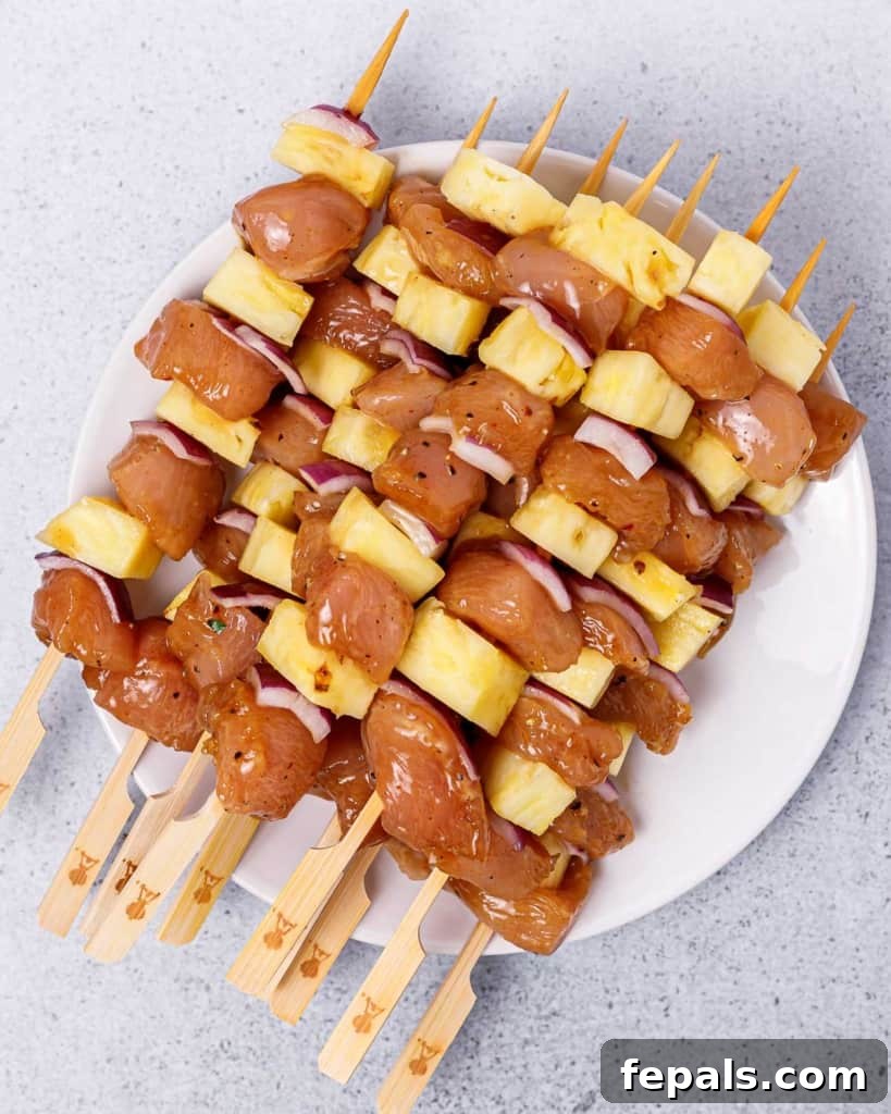 Grilled Umami Chicken Skewers 7 Uncooked chicken kabobs, perfectly threaded with pineapple and red onion, laid out on a white plate.