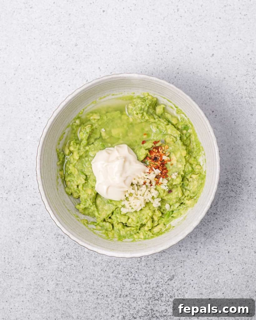 Adding mayonnaise, minced garlic, and chili flakes to the mashed avocado mixture in a bowl, preparing it for stirring.