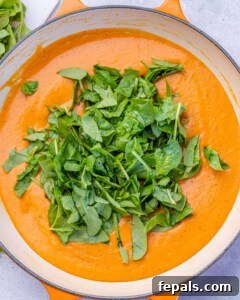 Chopped fresh baby spinach sitting on top of smooth red pepper sauce in a skillet, ready to stir in.