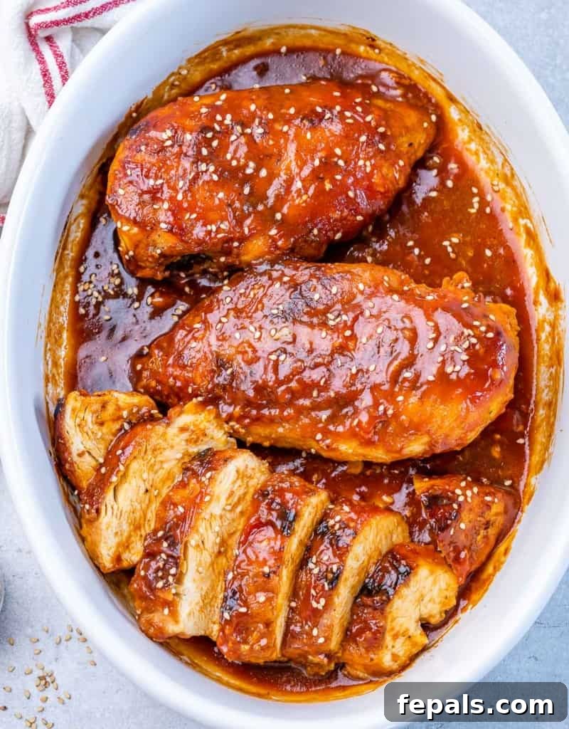 Sticky Honey Sriracha Chicken 11 Top view of three baked chicken breasts, one of which is sliced to show its juicy interior, garnished with sesame seeds in a white dish.